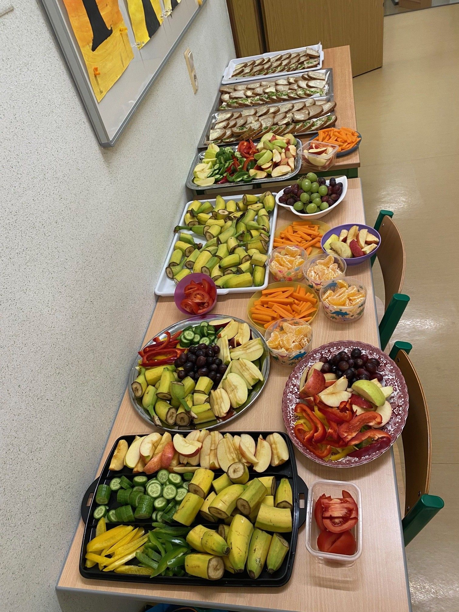 Ein langer Tisch mit verschiedenen Tellern von Obst und Gemüse, darunter Äpfel, Bananen, Karotten und Paprika. Es sind auch Schalen mit Trauben, Orangen und anderen Früchten vorhanden.