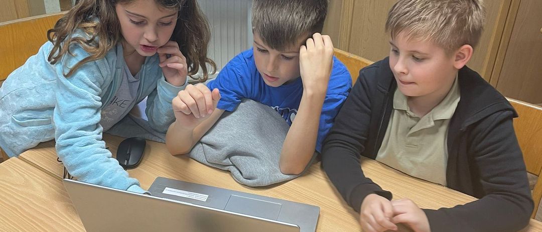 Drei Kinder sitzen an einem Tisch und konzentrieren sich auf einen Laptop. Ein Mädchen in hellblau schaut auf den Bildschirm, während zwei Jungen in blauer und schwarzer Jacke aufmerksam zusehen.