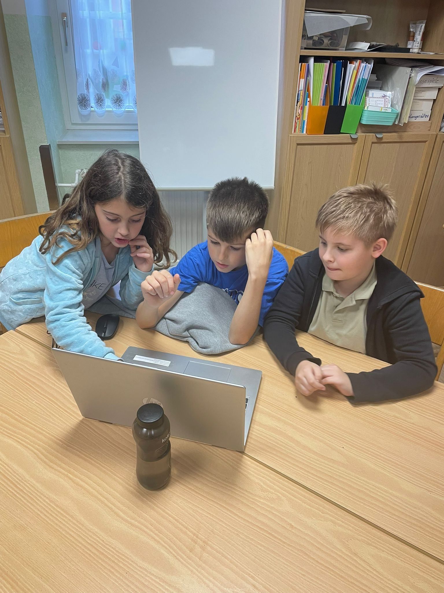 Drei Kinder sitzen an einem Schreibtisch und konzentrieren sich auf einen Laptop-Bildschirm. Ein Mädchen in Blau schaut aufmerksam, ein Junge in Blau stützt seinen Kopf auf seine Hand und ein anderer Junge in Schwarz lehnt sich vor.