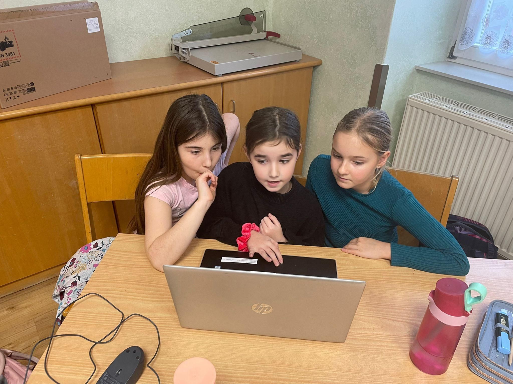 Drei Mädchen sitzen an einem Schreibtisch und schauen auf einen Laptop. Ein Mädchen hat ein rosafarbenes Band am Handgelenk.