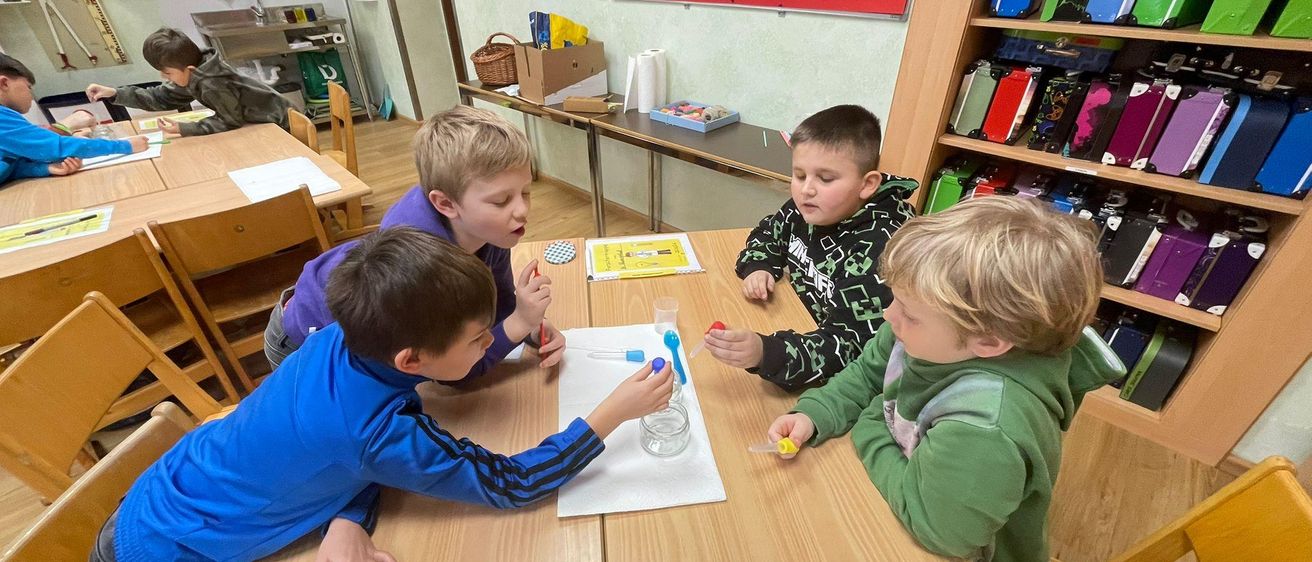Vier Jungen sind in einem Klassenzimmer mit einem wissenschaftlichen Experiment beschäftigt, wobei einer einen Pipettenhalter und der andere ein Reagenzglas hält.