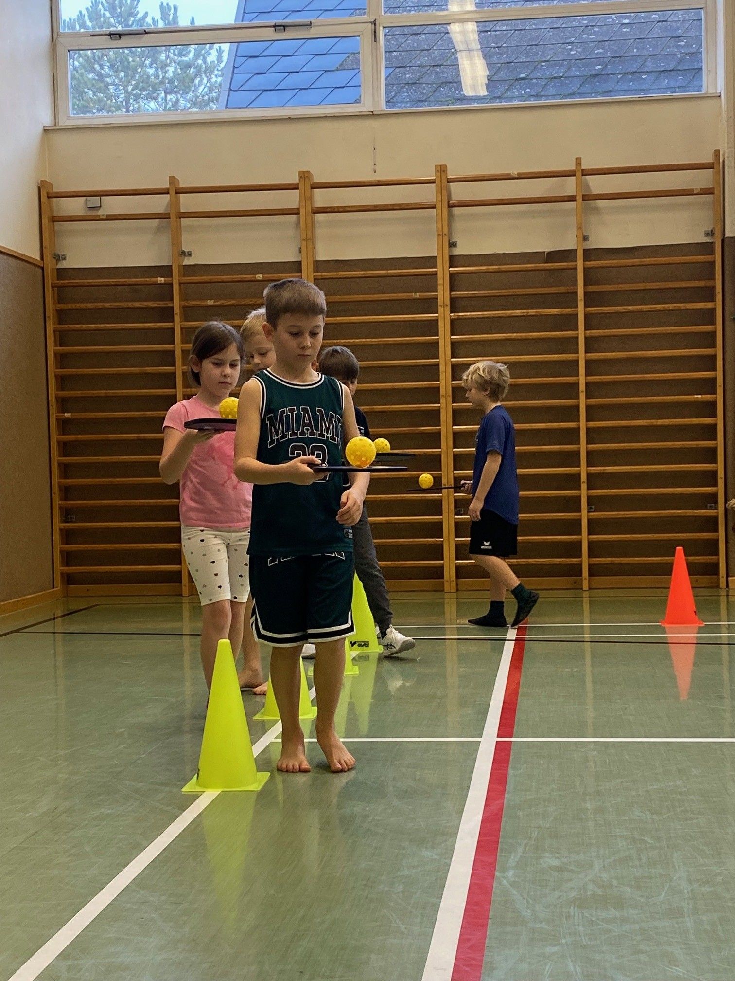 Eine Gruppe von Kindern spielt mit gelben Bällen in einer Turnhalle, wobei ein Junge in einem grünen Trikot einen Schläger hält.