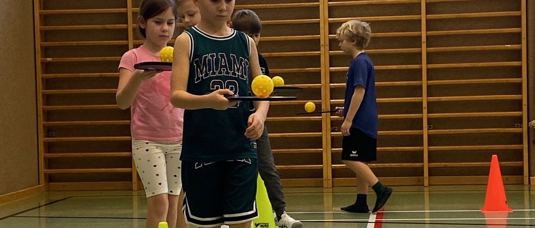 Kinder üben Ball-Handling-Fähigkeiten in einer Turnhalle mit Holzregalen im Hintergrund und gelben Kegeln auf dem Boden.