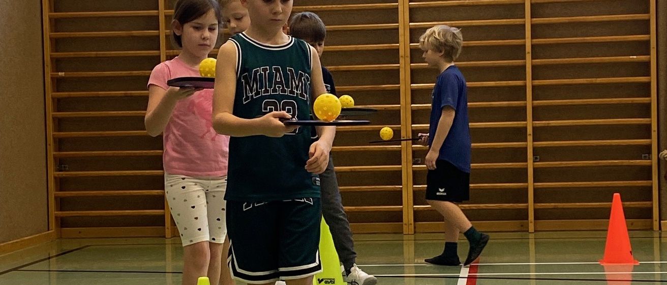 Kinder üben Ball-Handling-Fähigkeiten in einer Turnhalle mit Holzregalen im Hintergrund und gelben Kegeln auf dem Boden.