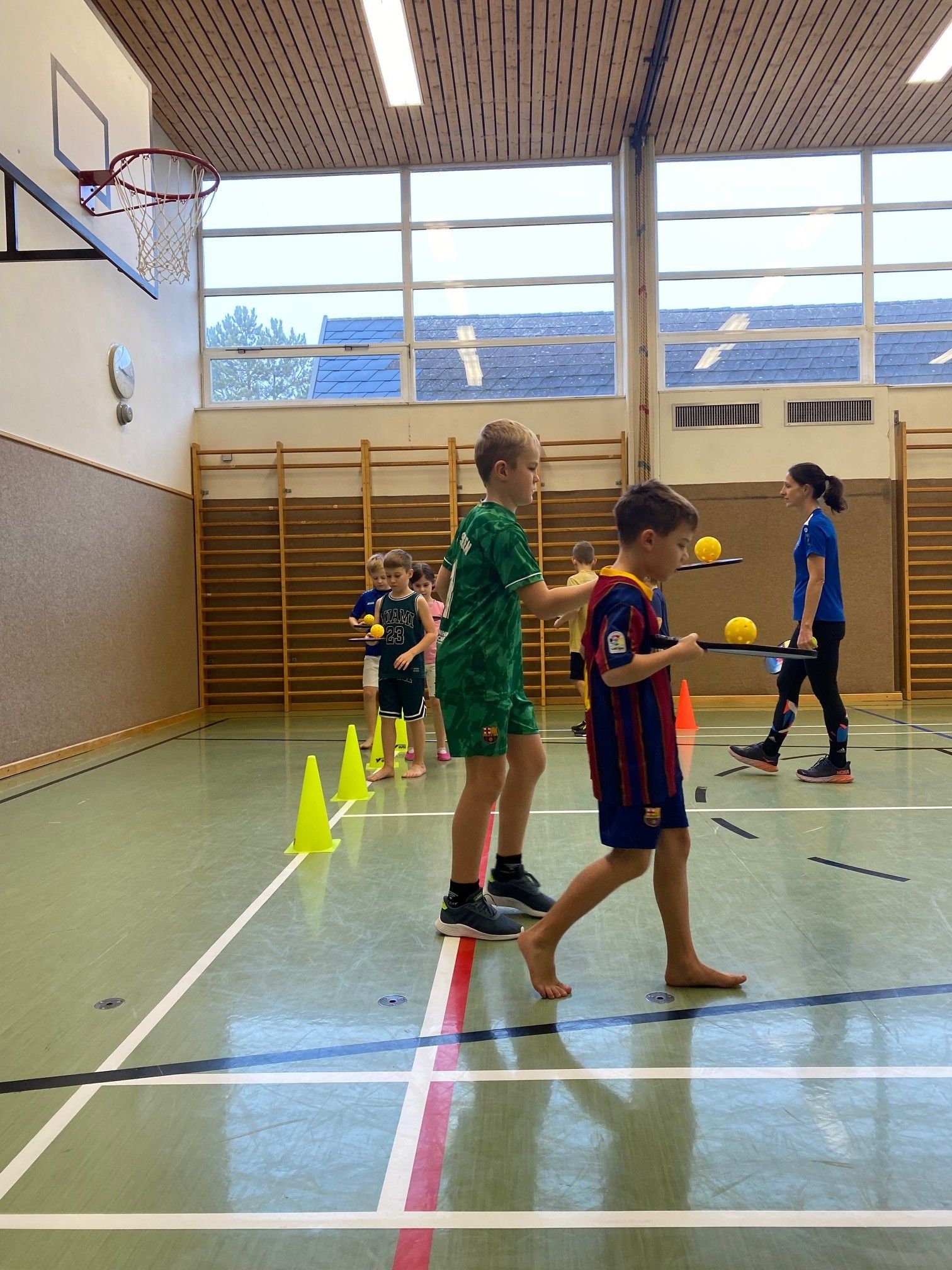 Kinder üben das Jonglieren mit gelben Bällen in einer Turnhalle, eine Frau beobachtet. Der Platz ist mit Linien und Kegeln markiert.