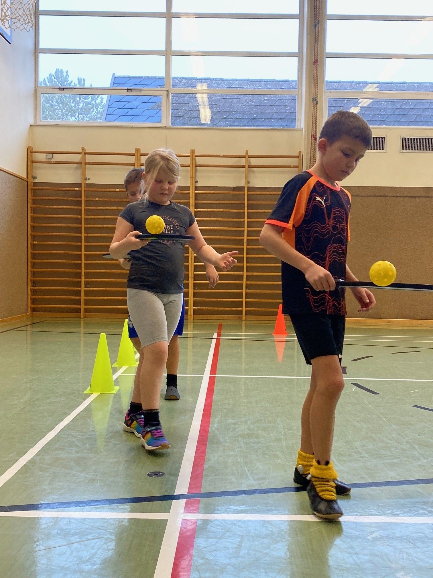 Zwei Kinder spielen mit gelben Bällen auf einem Spielfeld mit Kegeln und einer Holzstruktur im Hintergrund.