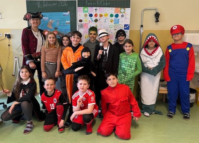 Eine Gruppe von Kindern in verschiedenen Kostümen, darunter ein Pirat, ein Hai und ein Mario-Charakter, posiert für ein Foto im Klassenzimmer.