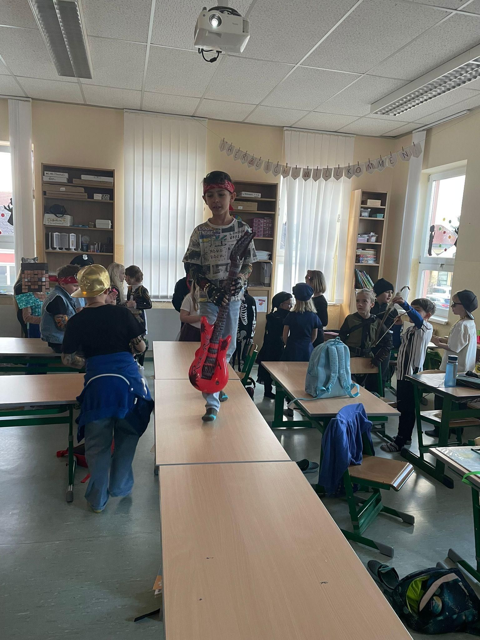 Ein Klassenzimmer mit Kindern in Kostümen, einer hält eine Gitarre, während andere zuschauen und einige an Tischen sitzen.