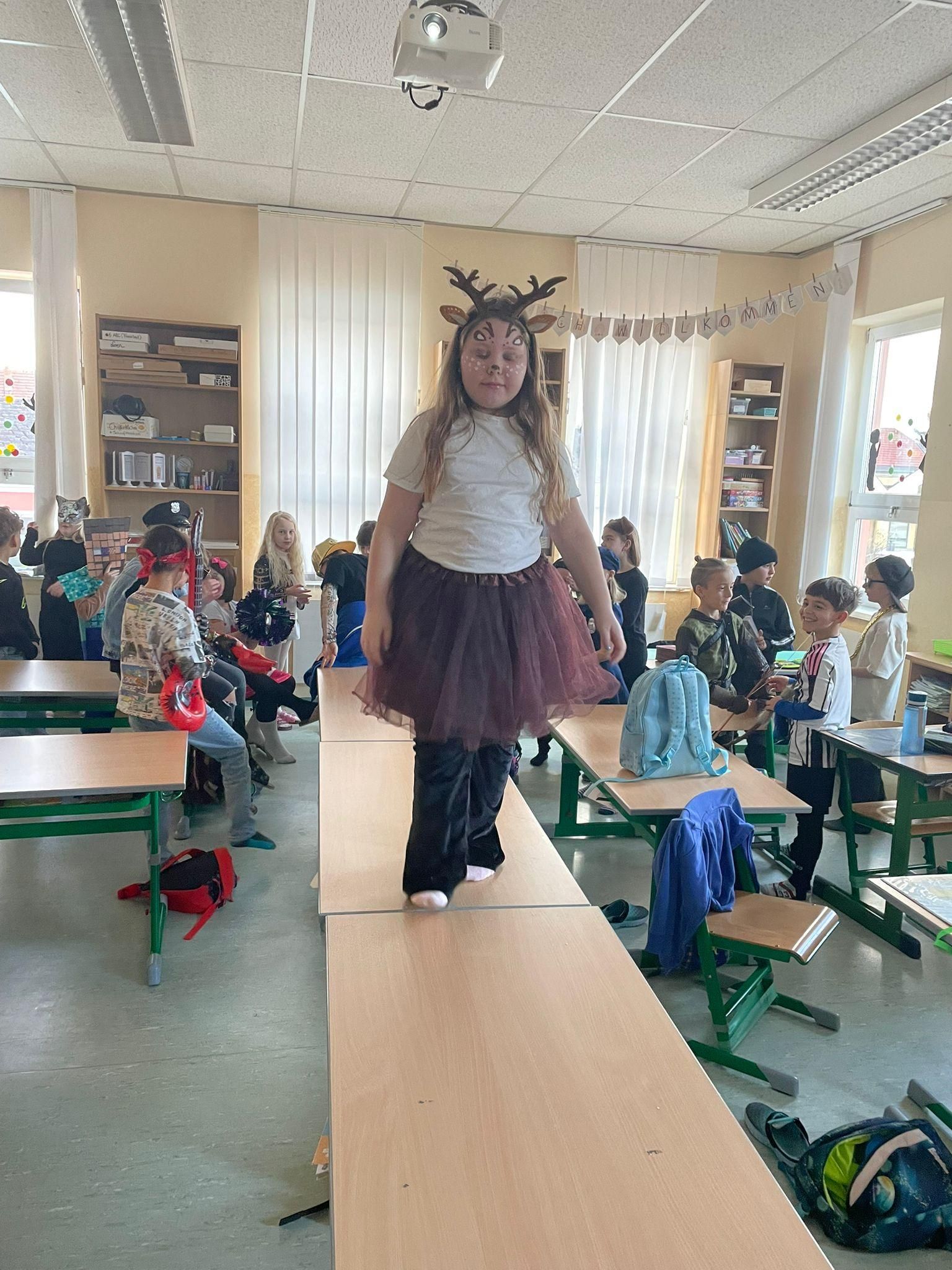 Ein junges Mädchen, verkleidet als Rentier, geht auf einer erhöhten Plattform in einem Klassenzimmer, umgeben von Kindern in Kostümen.