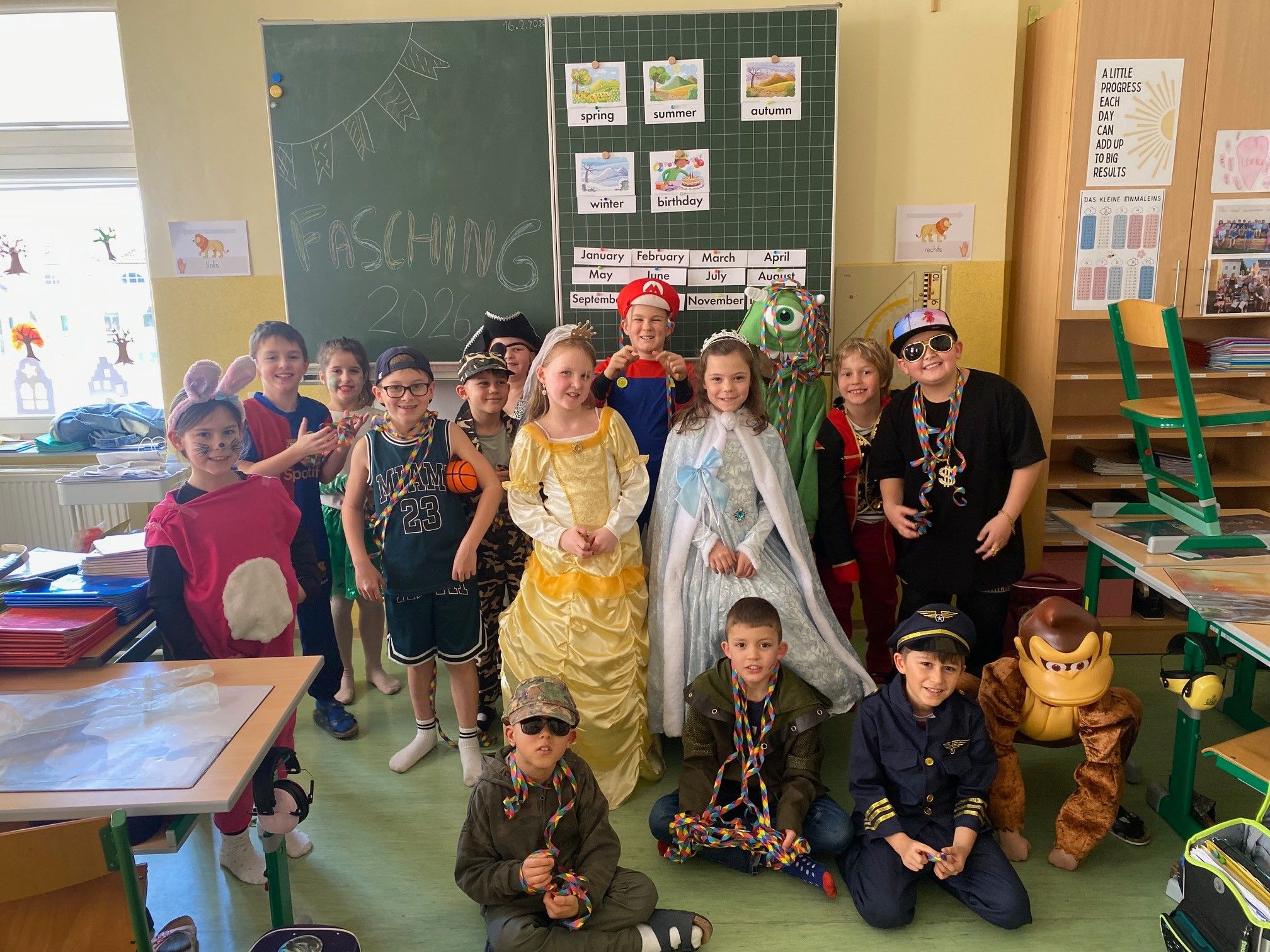 Ein Klassenzimmer voller Kinder in Kostümen, einige sitzend und einige stehend, mit einer Tafel im Hintergrund.