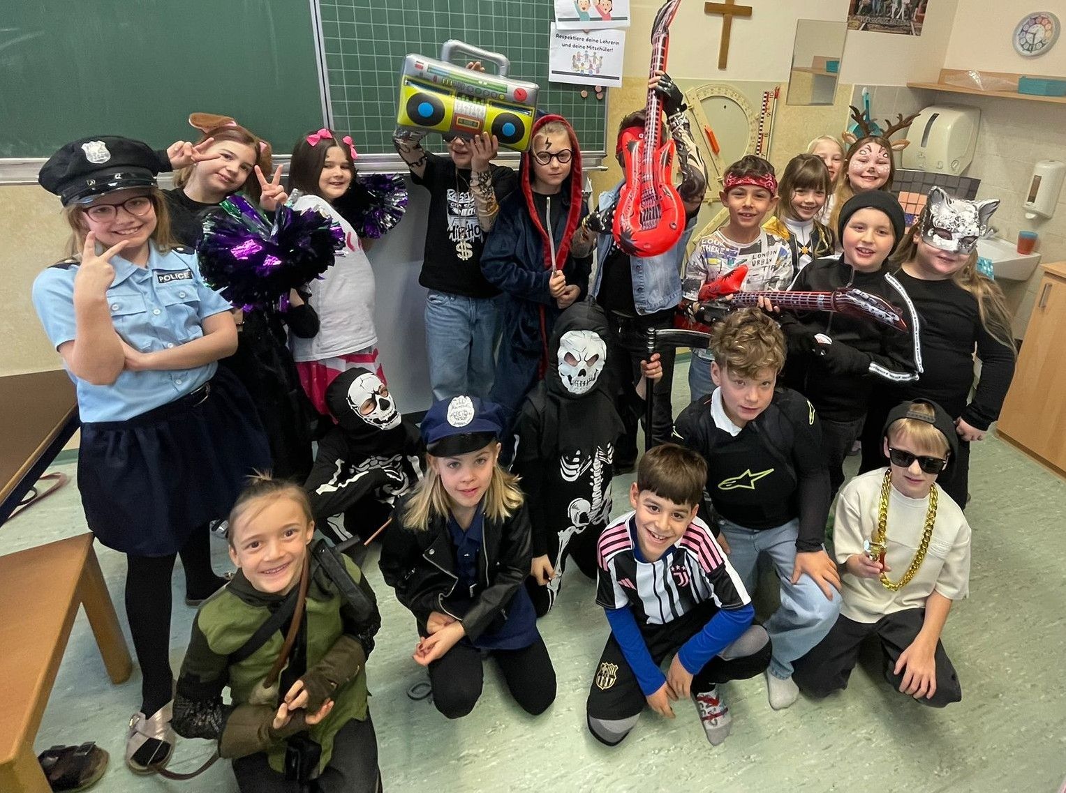 Eine Gruppe von Kindern in verschiedenen Kostümen, einige halten Requisiten wie Gitarren und Boomboxen, posieren für ein Foto im Klassenzimmer.