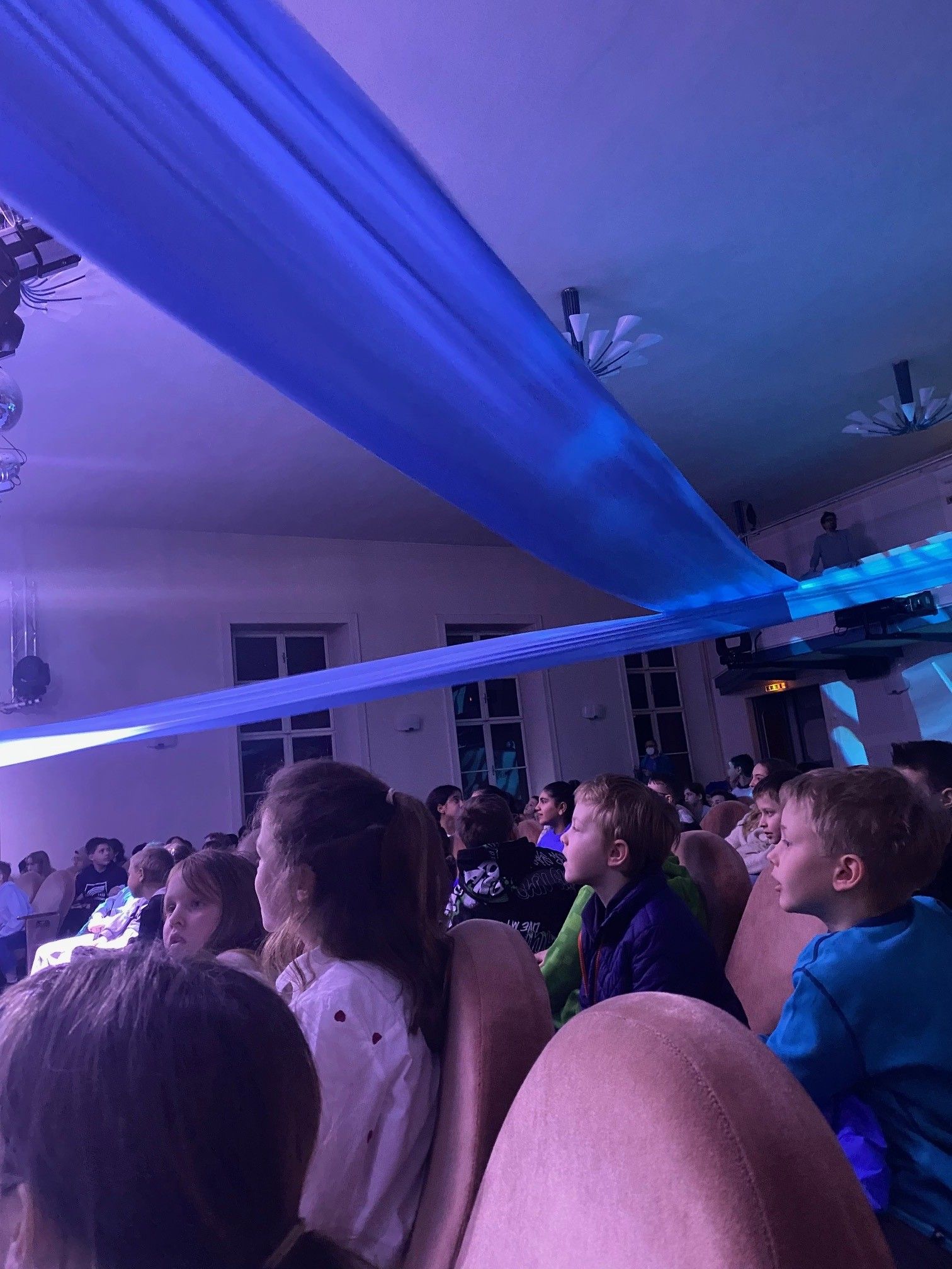 Eine Gruppe von Kindern und Erwachsenen sitzt in einem Raum mit blauer Beleuchtung und dekorativen Stoffen, die von der Decke hängen.
