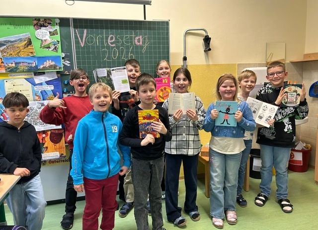 Eine Gruppe von Kindern in einem Klassenzimmer hält Bücher in der Hand und lächelt für ein Foto. Sie stehen vor einer Pinnwand mit verschiedenen Bildern und Texten.