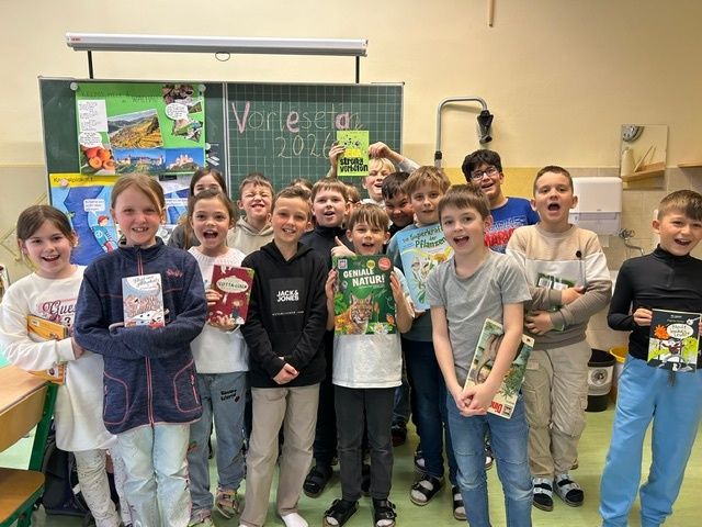 Eine Gruppe von Kindern in einem Klassenzimmer hält Bücher und lächelt für ein Foto. Sie stehen vor einer Tafel mit dem Text 'Vorleseta 2026'.