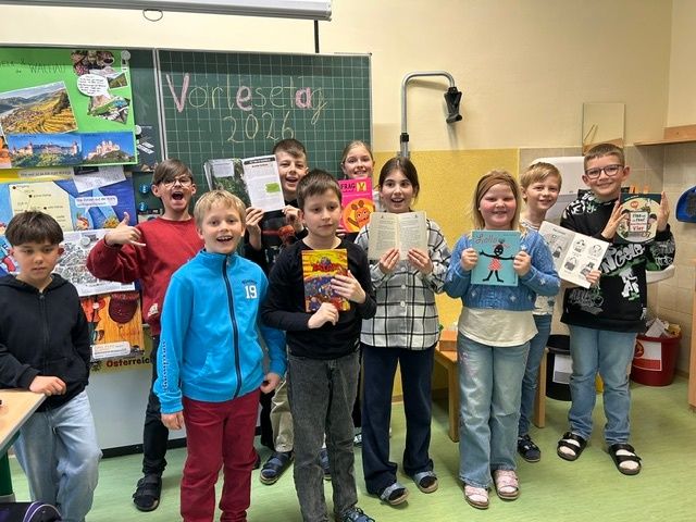 Eine Gruppe von Kindern im Klassenzimmer, die Bücher halten und lächeln, mit einer Tafel im Hintergrund.