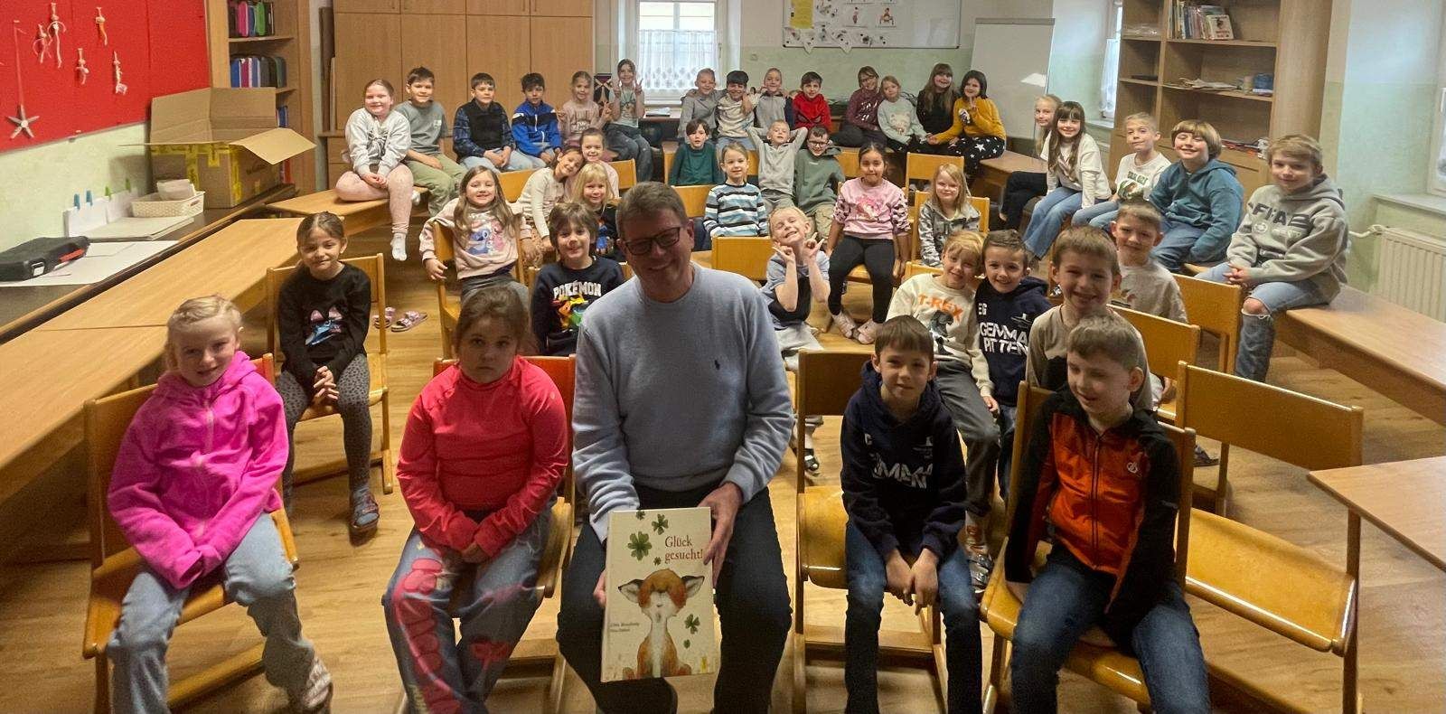 Eine Gruppe von Kindern und einem erwachsenen Mann sitzen in einem Klassenzimmer, lächeln und posieren für ein Foto. Der Mann hält ein Buch.