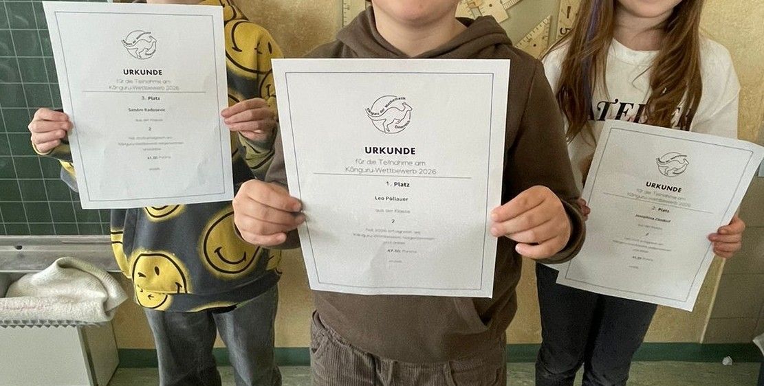 Drei Kinder stehen zusammen und halten Urkunden in der Hand, ein Junge in einer braunen Kapuzenjacke, ein anderer in einem gelben Shirt und ein Mädchen in einem weißen Shirt. Sie lächeln und posieren für ein Foto.