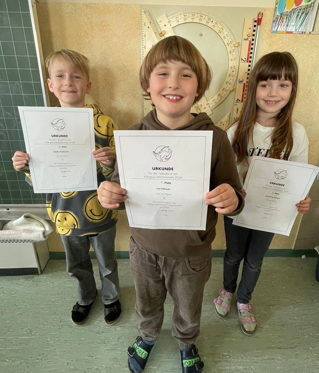 Drei Kinder, zwei Jungen und ein Mädchen, stehen zusammen und halten Urkunden in den Händen. Der Junge in der Mitte lächelt, während die anderen in die Kamera schauen. Sie befinden sich in einem Raum mit einer grünen Wand und einem weißen Regal.