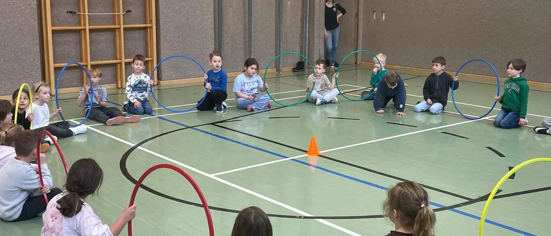 Kinder spielen mit Hula-Hoops in einer Turnhalle, einige sitzen auf dem Boden und andere stehen. Eine Frau steht in der Nähe der Tür.