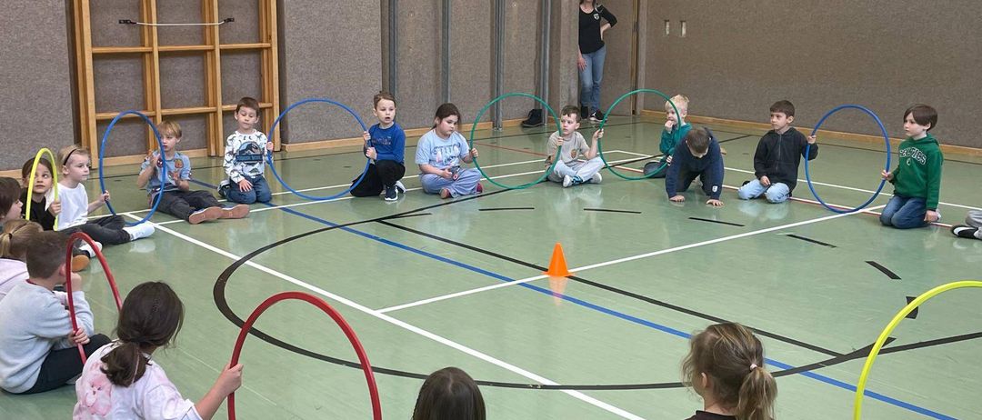 Kinder spielen mit Hula-Hoops in einer Turnhalle, einige sitzen auf dem Boden und andere stehen. Eine Frau steht in der Nähe der Tür.