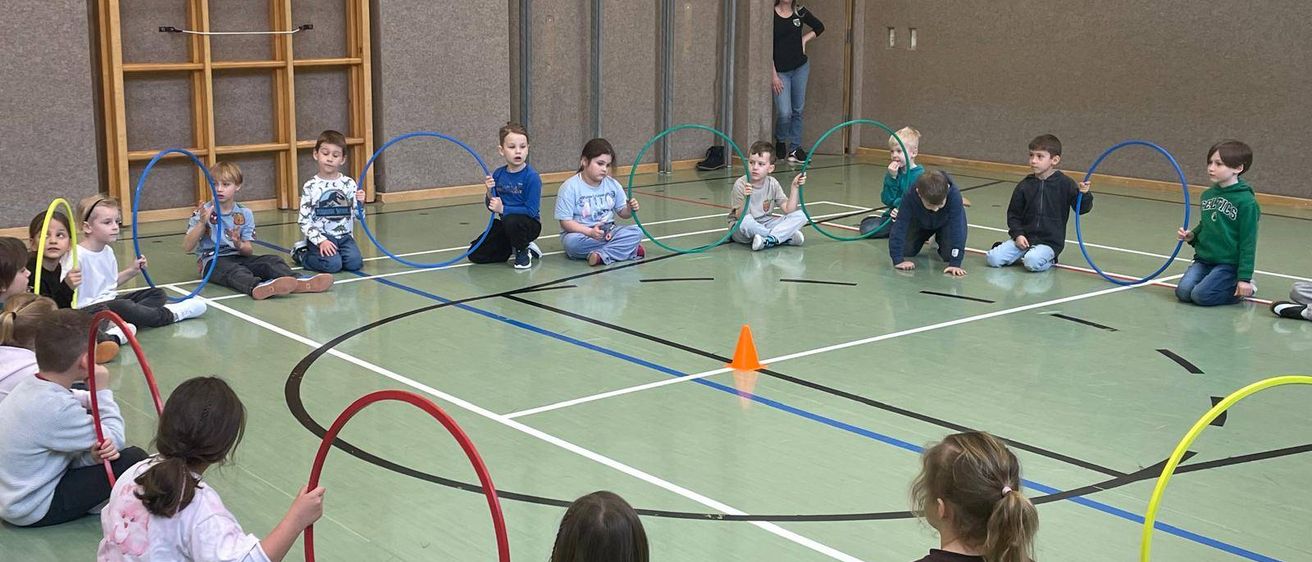Kinder spielen mit Hula-Hoops in einer Turnhalle, einige sitzen auf dem Boden und andere stehen. Eine Frau steht in der Nähe der Tür.