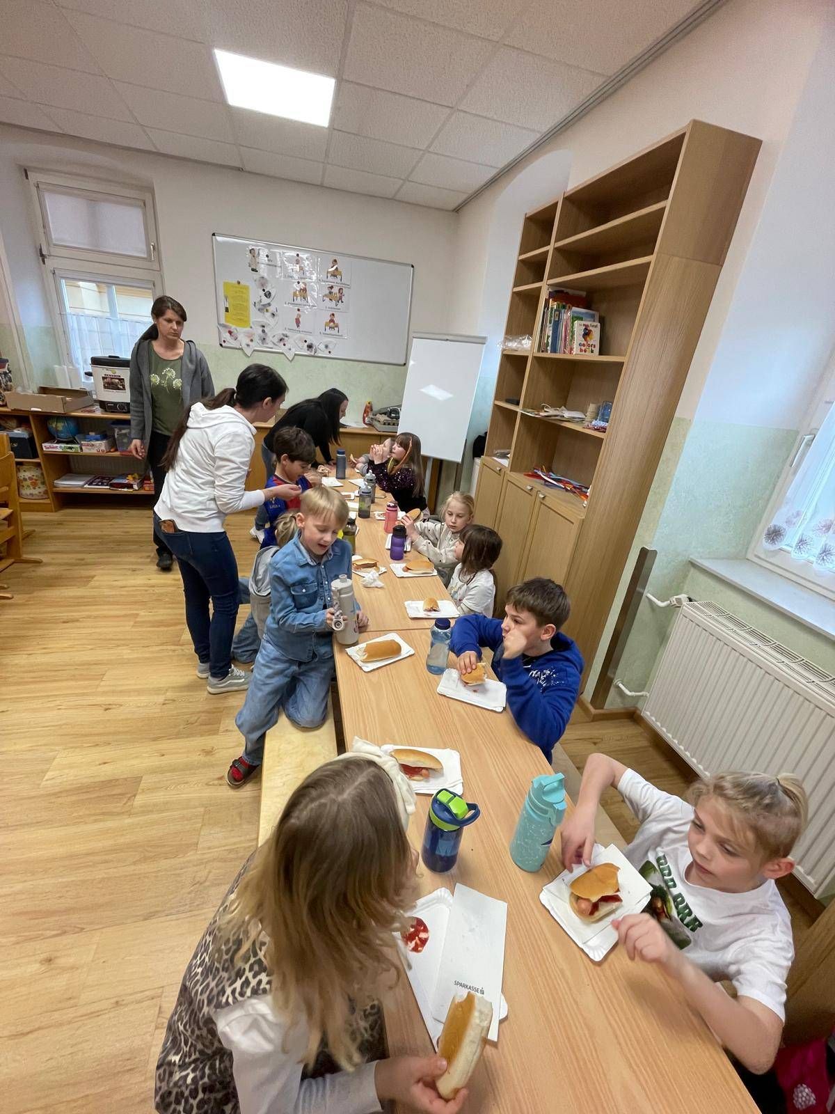 Eine Gruppe von Kindern und Erwachsenen sitzt an einem langen Tisch in einem Klassenzimmer, isst Sandwiches und trinkt aus Flaschen. Eine Frau in einer grauen Kapuzenjacke steht hinter dem Tisch, und eine Tafel ist an der Wand.