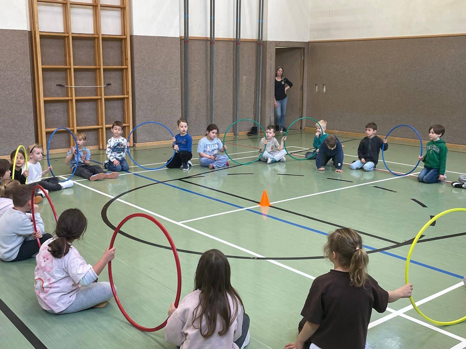 Eine Gruppe von Kindern sitzt im Kreis auf dem Gymnastikboden, hält Hula-Hoops und spielt ein Spiel. Eine Frau steht in der Nähe.