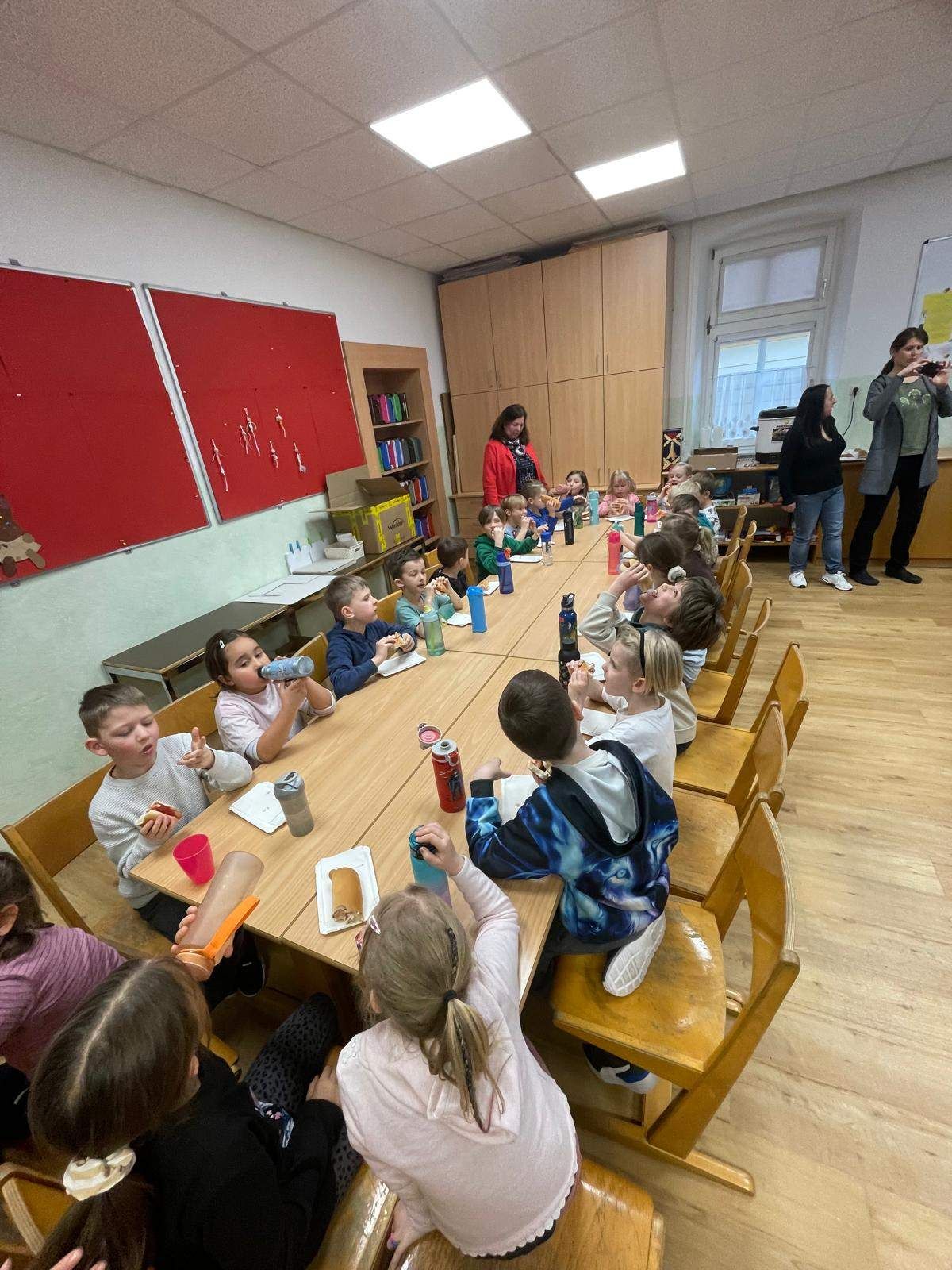 Eine Gruppe von Kindern sitzt an einem langen Tisch in einem Klassenzimmer, isst und trinkt. Eine Frau steht hinter ihnen, und eine andere Frau macht ein Foto.