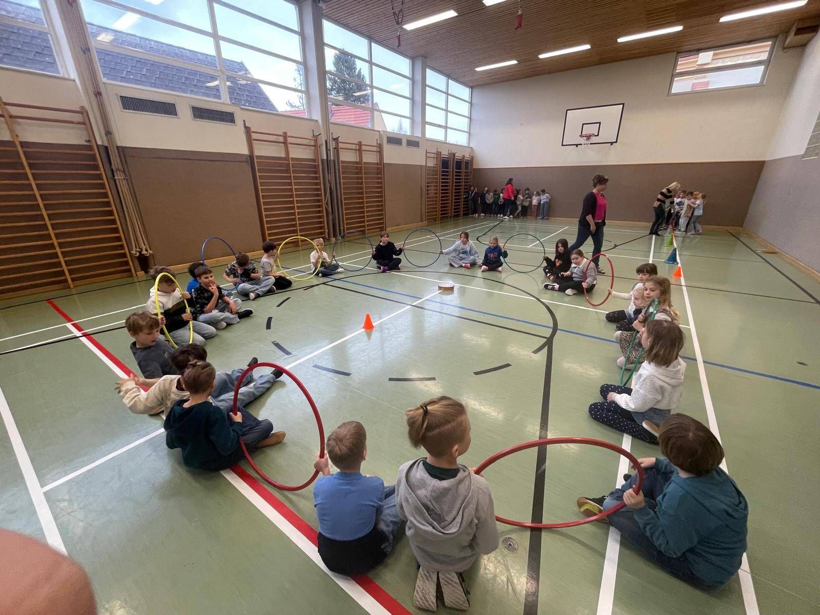 Kinder sitzen im Kreis auf dem Gymnastikboden und halten Hula-Hoops, eine Erwachsene steht in der Nähe. Die Turnhalle hat ein Basketballkorb, Fenster und eine Gruppe von Kindern im Hintergrund.