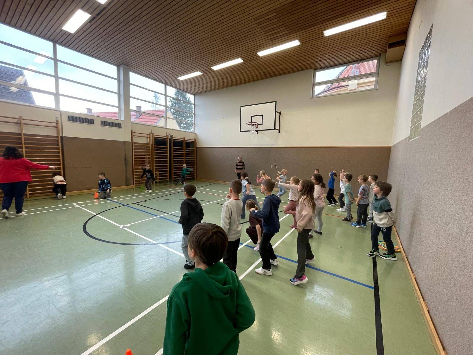 Kinder in einer Turnhalle, einige spielen mit orangefarbenen Kegeln, andere beobachten eine Person in einem schwarzen Shirt. Ein Basketballkorb ist an der Wand zu sehen.