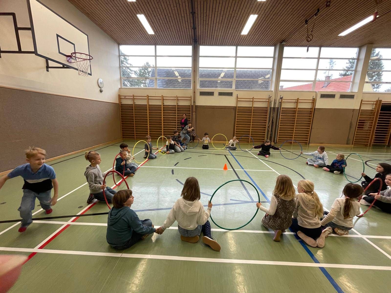 Kinder in einer Turnhalle, einige sitzend und andere stehend, beschäftigen sich mit einem Hula-Hoop-Aktivität. Der Raum ist gut beleuchtet mit großen Fenstern und einem sichtbaren Basketballkorb.
