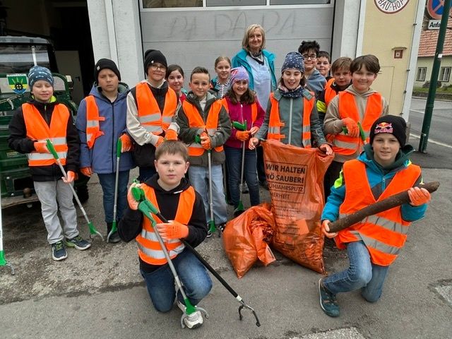 Eine Gruppe von Kindern in orangefarbenen Westen, die Reinigungswerkzeuge halten und vor einem Gebäude mit einer Frau stehen. Sie nehmen an einer Gemeinschaftsreinigungsaktion teil.