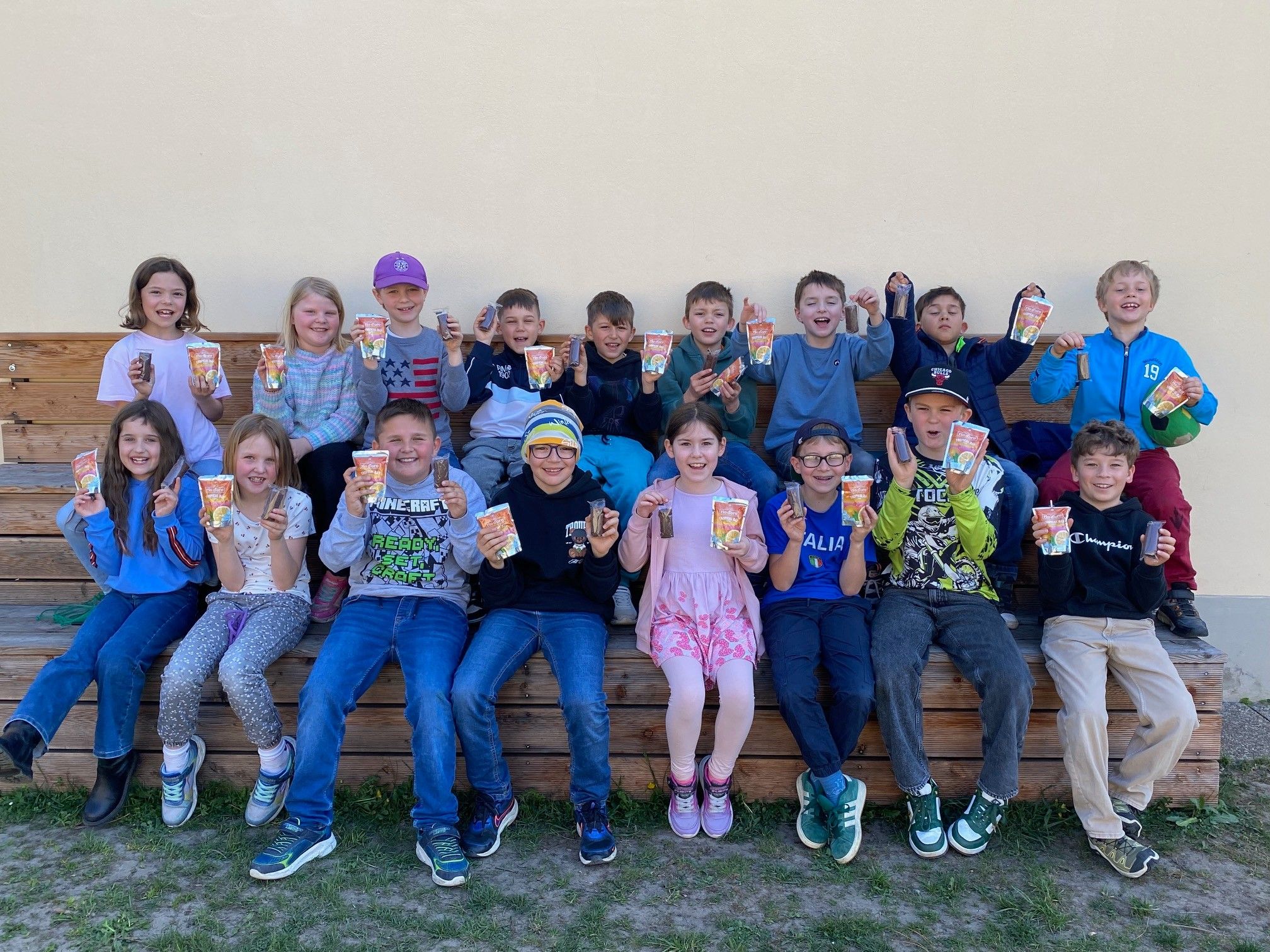 Eine Gruppe von Kindern sitzt auf einer Bank, hält kleine Tüten mit Snacks hoch und posiert für ein Foto.