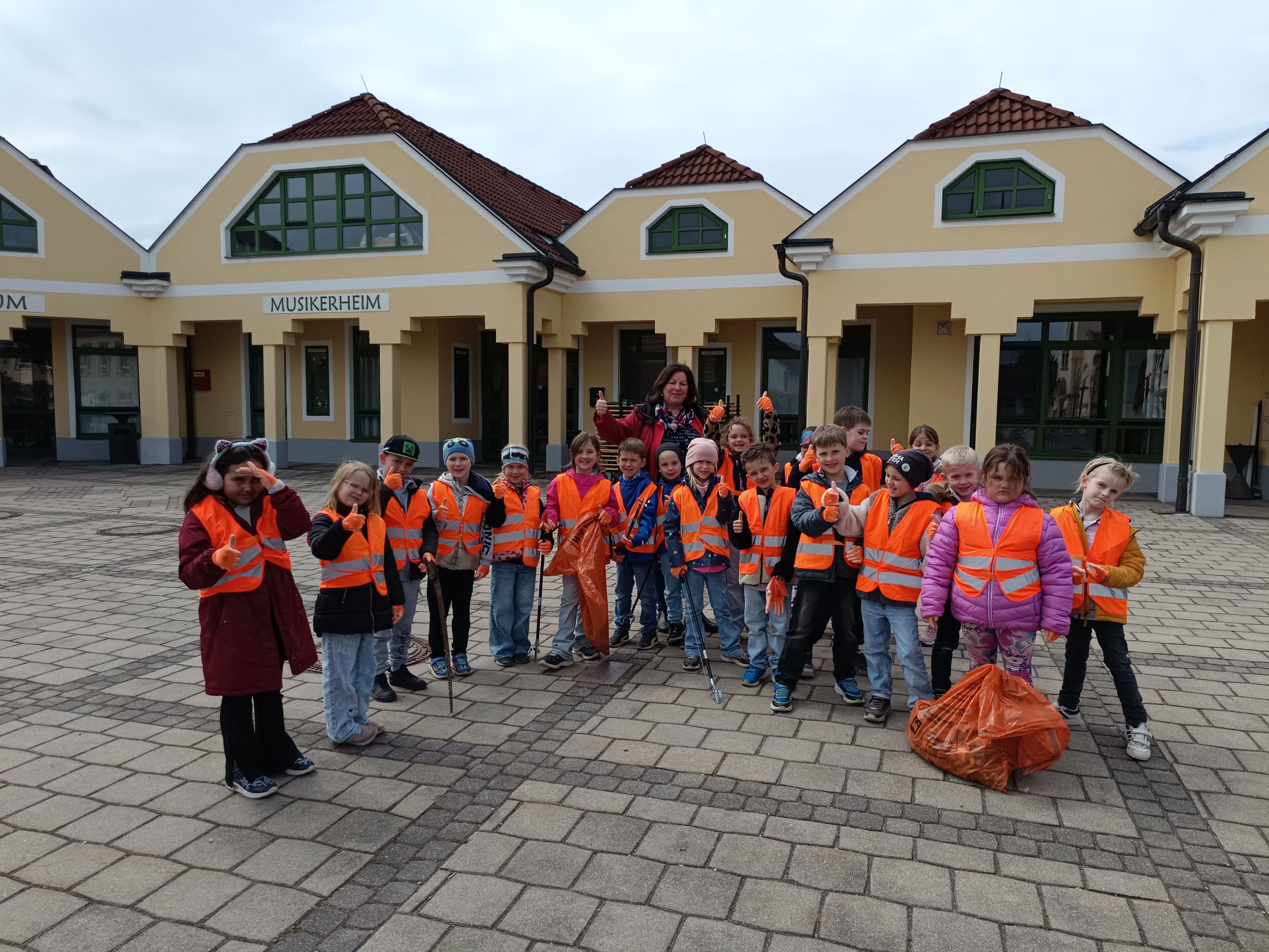 Eine Gruppe von Kindern in orangefarbenen Westen, die Mülltüten halten, steht vor einem Gebäude. Sie lächeln und posieren für ein Foto.