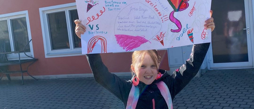 Ein junges Mädchen hält ein farbenfrohes Schild mit der Aufschrift 'Los Breitenau!' vor einem Gebäude. Das Schild enthält auch Illustrationen und Text in mehreren Sprachen.