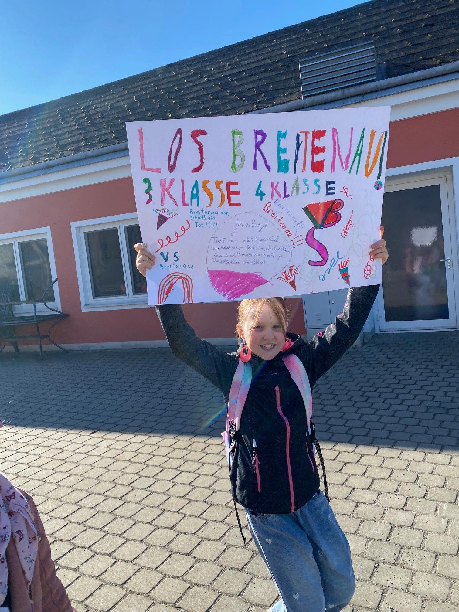 Ein kleines Mädchen hält ein buntes Schild mit den Worten 'Los Breitenau' und '3 Klasse 4 Klasse' vor einem Gebäude.