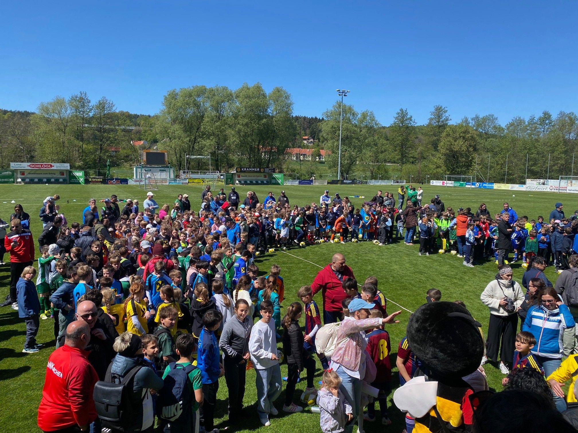 Eine große Gruppe von Kindern und Erwachsenen versammelt sich auf einem Rasenfeld, wahrscheinlich für eine Sportveranstaltung. Einige tragen Trikots mit Nummern, andere sind in Freizeitkleidung. Ein Mann in einem roten Jackett zeigt auf etwas. Im Hintergrund befinden sich Bäume, eine Anzeigetafel und ein Gebäude.