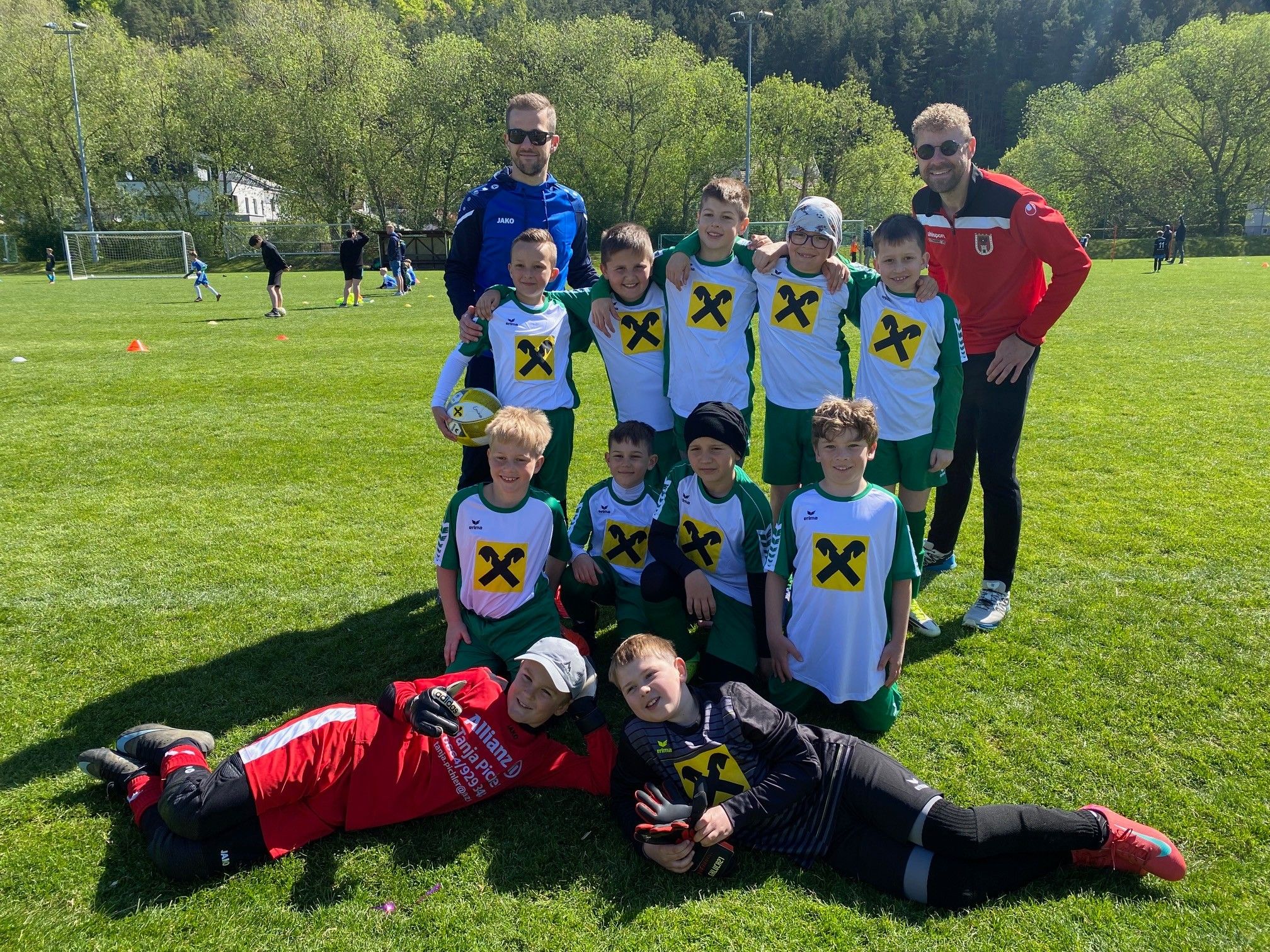 Eine Gruppe junger Fußballspieler in grünen und weißen Trikots mit gelben X's posiert für ein Foto mit zwei Trainern an einem sonnigen Tag.