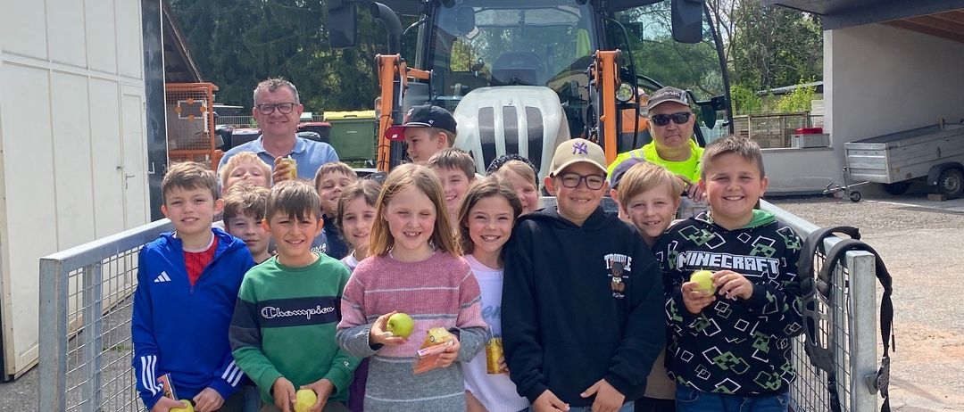 Eine Gruppe von Kindern und Erwachsenen steht im Heck eines Traktors und hält Äpfel in der Hand. Sie lächeln und posieren für ein Foto.