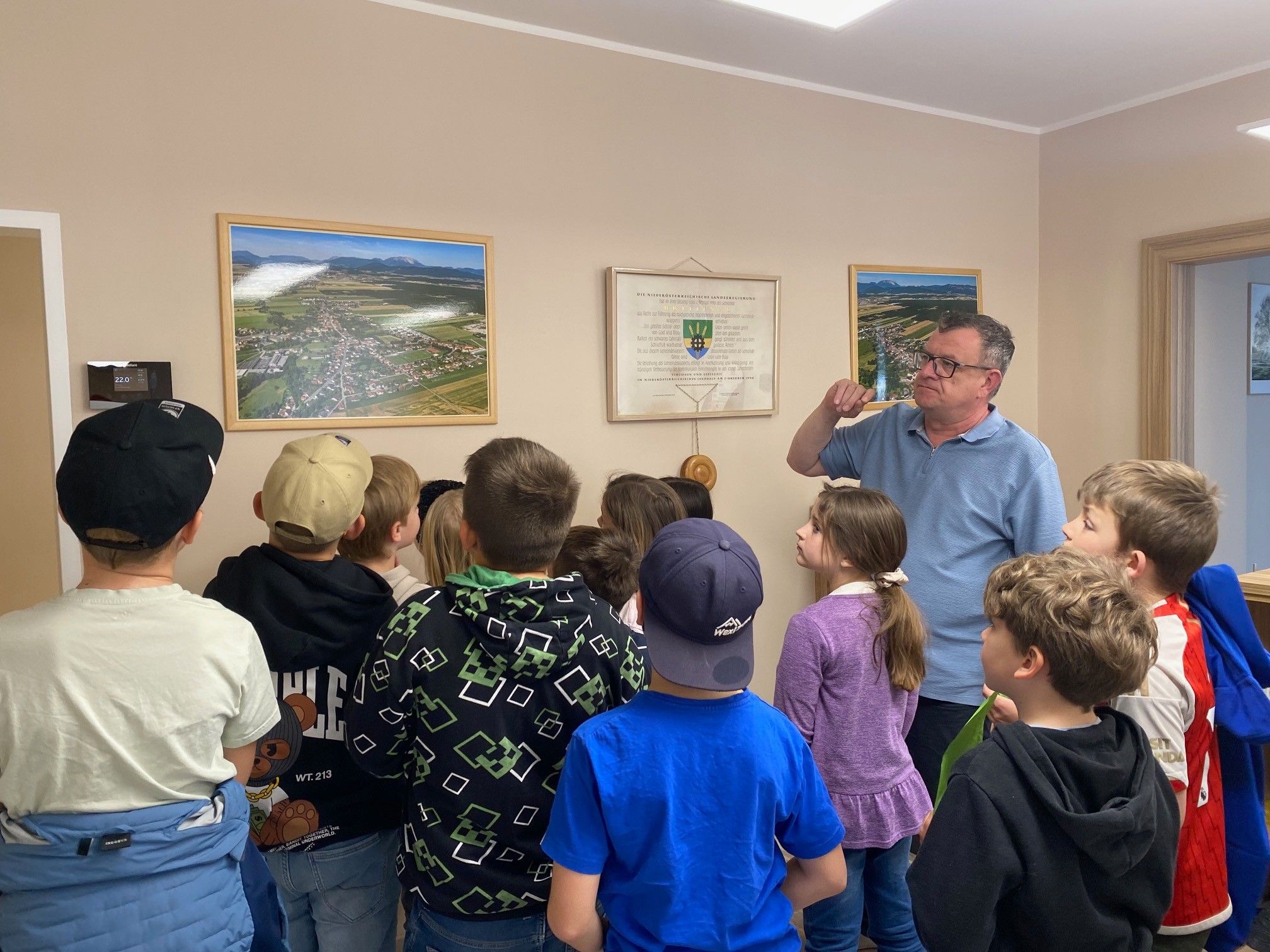 Eine Gruppe von Kindern und einem Mann versammeln sich in einem Raum und betrachten ein gerahmtes Bild an der Wand. Der Mann erklärt ihnen etwas.