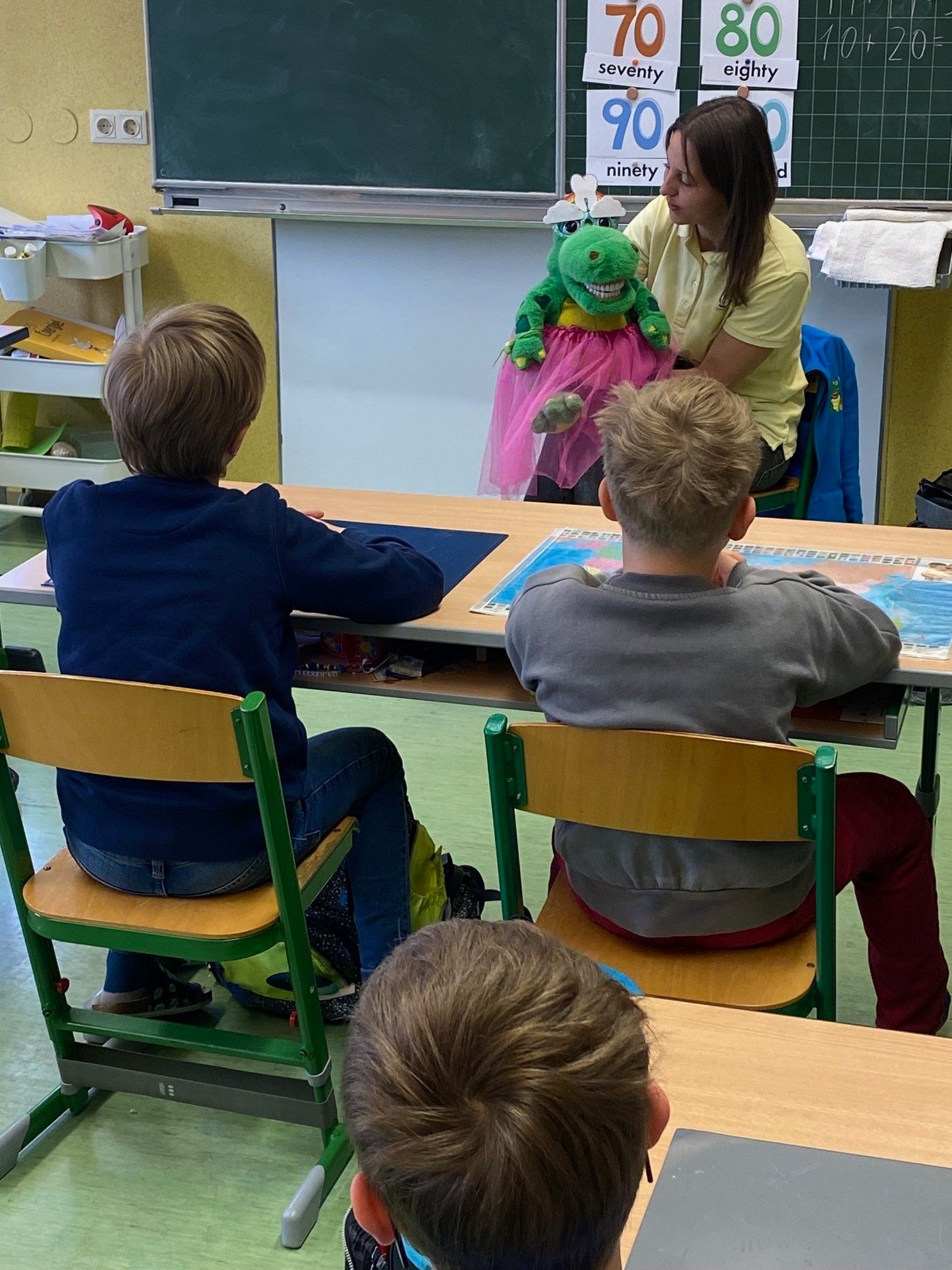 Eine Frau mit einer Puppe sitzt hinter zwei Jungen an einem Schreibtisch im Klassenzimmer.