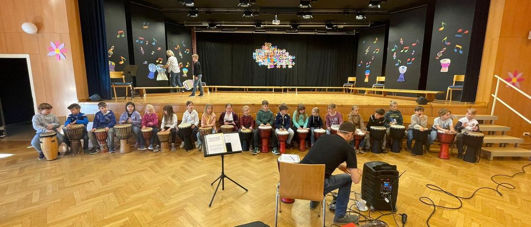 Eine Gruppe von Kindern sitzt mit Trommeln auf dem Boden, vor einem Mann, der sitzt und eine Gitarre spielt. Der Raum hat eine Bühne mit Noten an den Wänden und einem Holzboden.