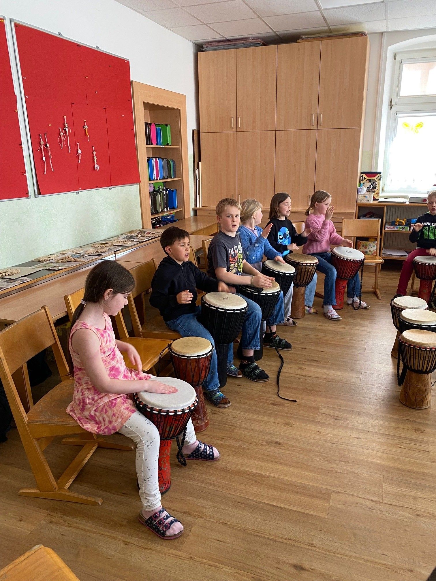 Eine Gruppe von Kindern sitzt in einem Klassenzimmer, jedes spielt eine Trommel. Sie tragen Freizeitkleidung und scheinen an einer Musikaktivität beteiligt zu sein.