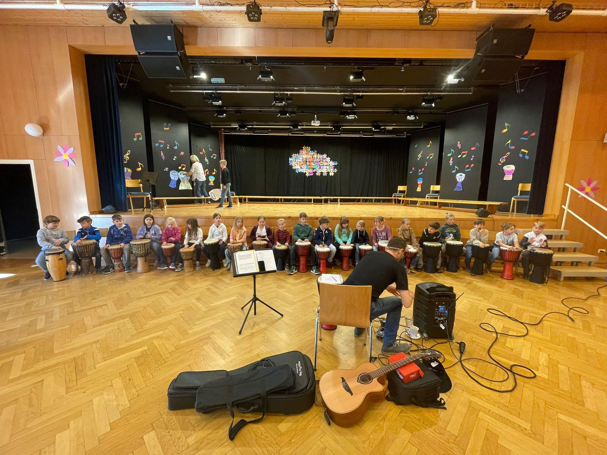 Eine Gruppe von Kindern sitzt auf dem Boden und hält Trommeln, während ein Mann auf einem Stuhl vor ihnen sitzt und eine Gitarre spielt. Der Raum hat einen Holzboden und Wände mit musikalischen Noten.
