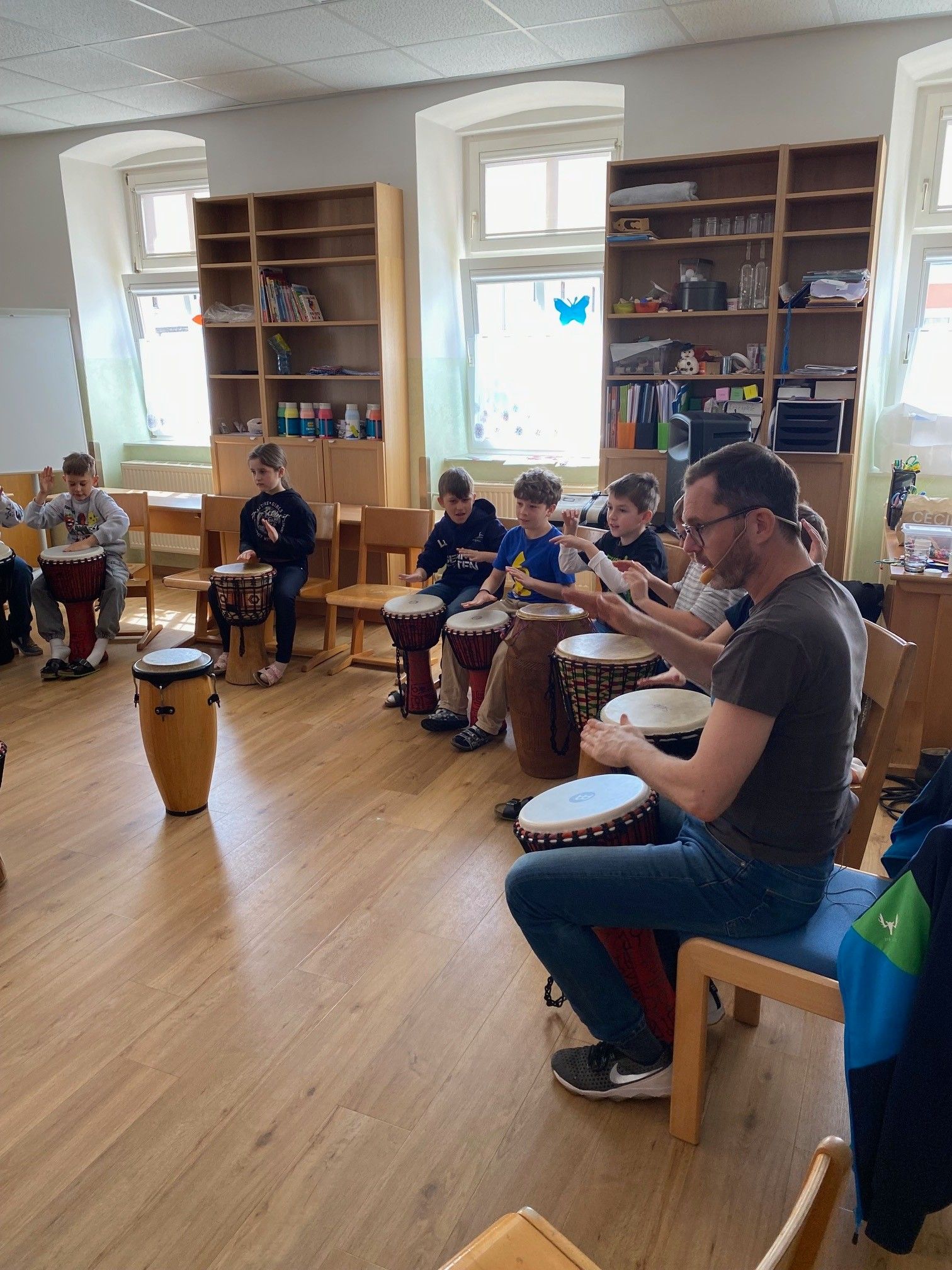 Eine Gruppe von Kindern spielt Trommeln in einem Klassenzimmer, angeleitet von einem Mann, der auf einem Stuhl sitzt.