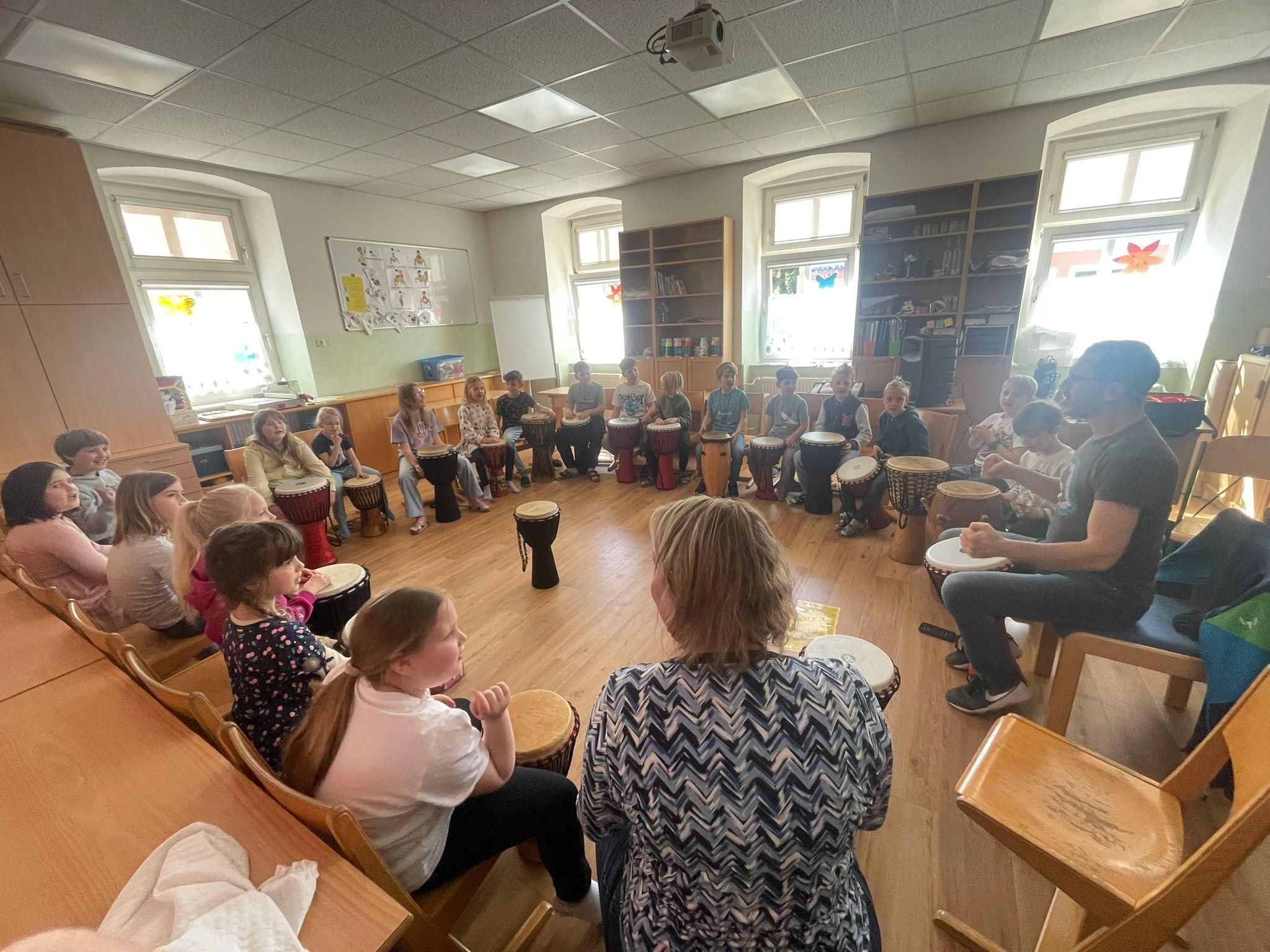 Eine Gruppe von Kindern und Erwachsenen versammelt sich in einem Raum und spielt Trommeln. Sie sitzen im Kreis um den Raum herum, wobei einige Erwachsene beobachten.