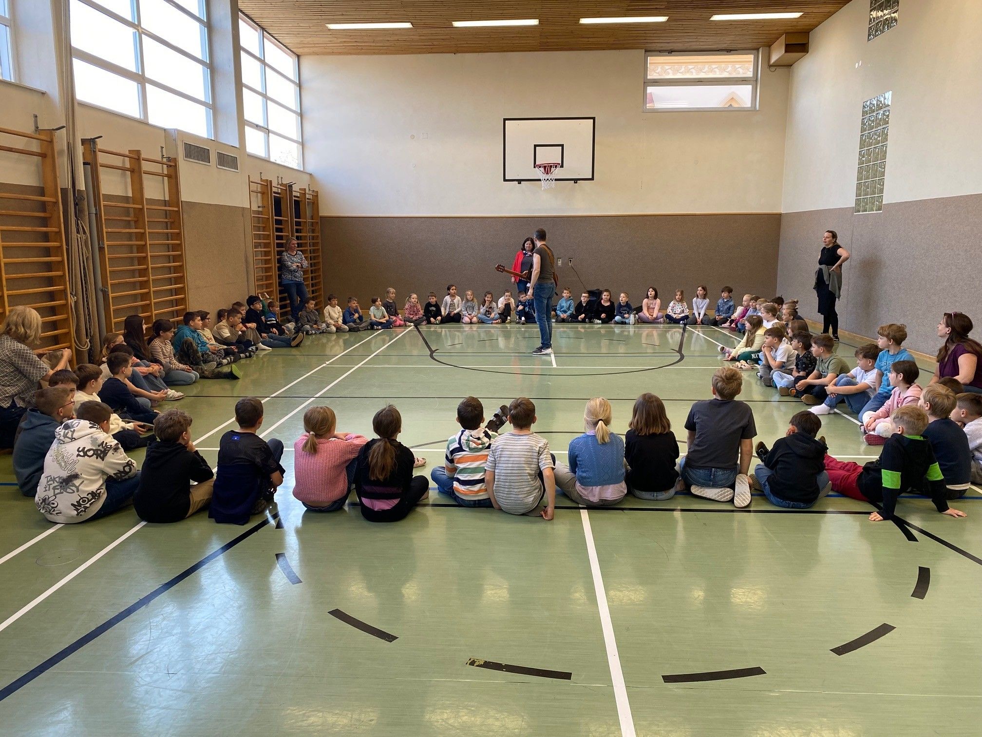 Eine Gruppe von Kindern und Erwachsenen versammelt sich in einer Turnhalle und beobachtet einen Mann, der Gitarre spielt. Die Kinder sitzen auf dem Boden, während die Erwachsenen in der Nähe des Basketballfeldes stehen.