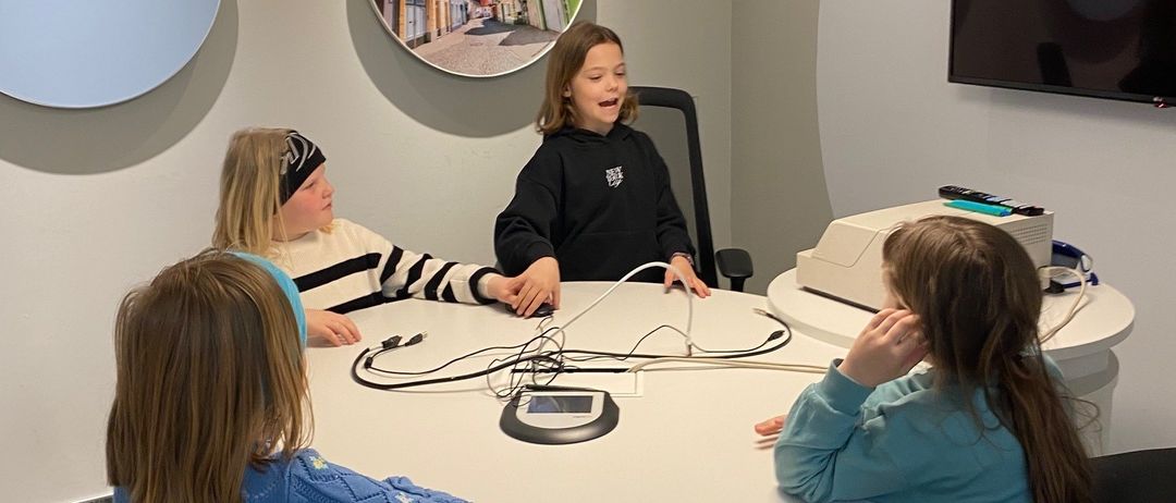 Vier junge Mädchen sitzen um einen runden Tisch in einem modernen Raum. Ein Mädchen trägt Kopfhörer und scheint zu sprechen. Ein Monitor und ein runder Bilderrahmen hängen an der Wand hinter ihnen.