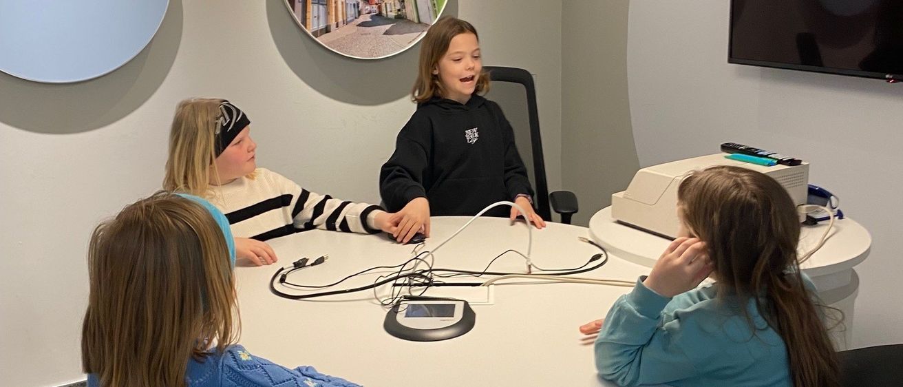 Vier junge Mädchen sitzen um einen runden Tisch in einem modernen Raum. Ein Mädchen trägt Kopfhörer und scheint zu sprechen. Ein Monitor und ein runder Bilderrahmen hängen an der Wand hinter ihnen.