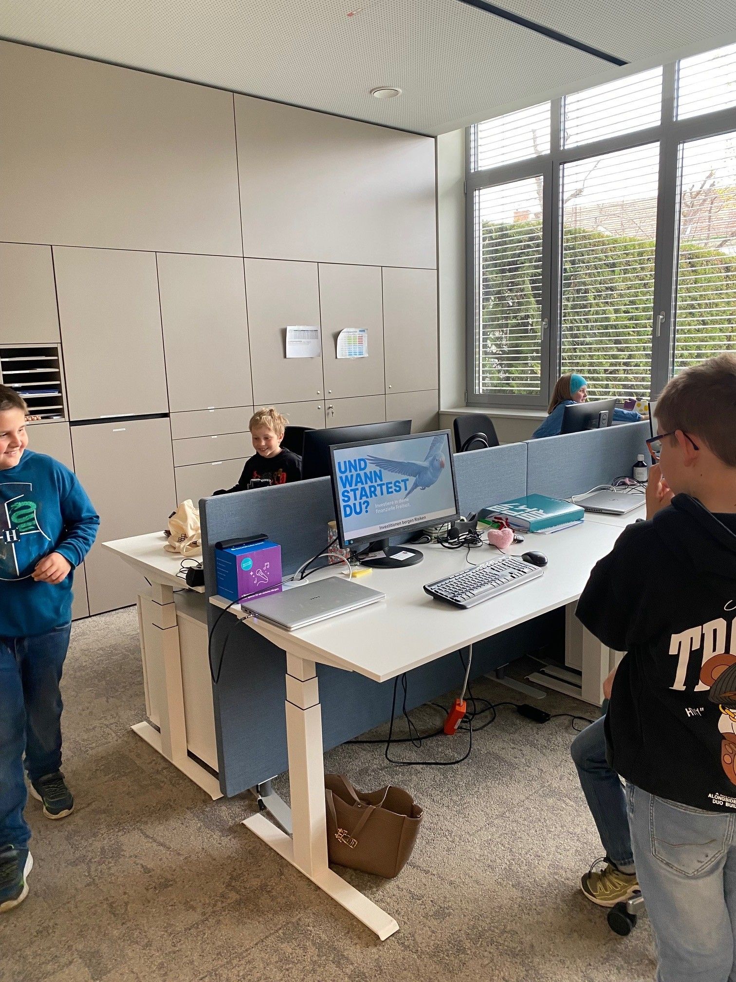 Drei Kinder sind in einer Büroumgebung. Ein Kind sitzt an einem Schreibtisch mit einem Computer, während die anderen beiden in der Nähe stehen. Der Schreibtisch hat verschiedene Gegenstände wie Tastatur, Maus und einen Monitor, der einen blauen Vogel zeigt.